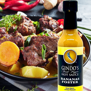 Bottle of Gindo's Fresh Pepper Hot Sauce Bananas Foster in front of a bowl of stew with meat and vegetables.