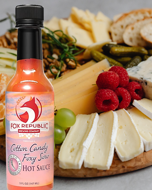 A bottle of Fox Republic Cotton Candy Foxy Sav Hot Sauce in front of a charcuterie board with cheese, raspberries, grapes, and bread.