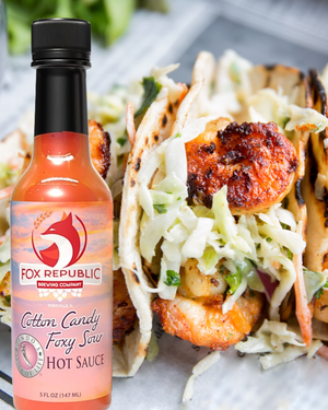 A bottle of Fox Republic Cotton Candy Foxy Saw Hot Sauce placed in front of grilled shrimp tacos filled with shredded cabbage on a light surface.