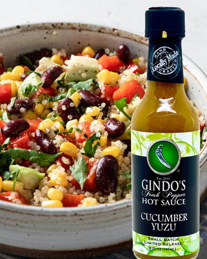 A bottle of Gindo's Fresh Pepper Hot Sauce Cucumber Yuzu sauce shown next to a close-up of a colorful quinoa salad containing red bell peppers, black beans, corn, and avocado in a speckled ceramic bowl.
