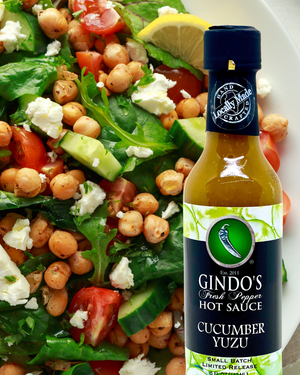 A bottle of Gindo's Fresh Pepper Hot Sauce Cucumber Yuzu sauce placed on the right side of a fresh salad with chickpeas, tomatoes, cucumbers, feta cheese, and a lemon wedge on the left.