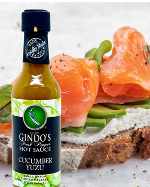 A bottle of Gindo's Fresh Pepper Hot Sauce, Cucumber Yuzu flavor, shown in the foreground with smoked salmon, avocado, and a slice of toast blurred in the background.