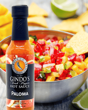 A bottle of Gindo's Paloma Fresh Pepper Hot Sauce in front of a bowl of colorful salsa with tortilla chips and lime wedges on a gray surface.