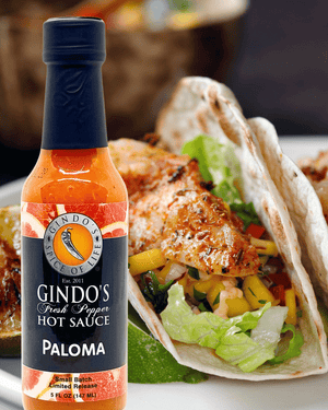 A bottle of Gindo's Paloma Fresh Pepper Hot Sauce placed in front of a taco filled with grilled chicken, lettuce, and peppers on a white plate.