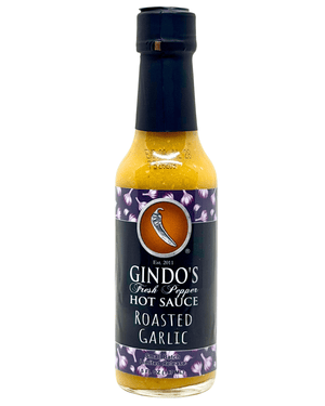 A bottle of Gindo's Fresh Peppers Roasted Garlic Hot Sauce on a plain white background. The bottle contains a yellow-orange sauce and features a black cap and a dark label with purple floral accents.