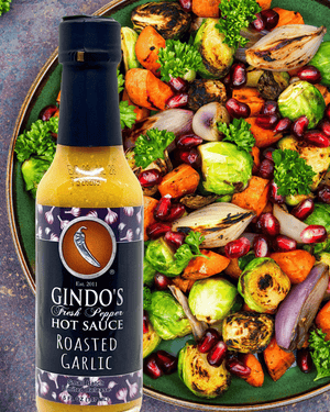 A bottle of Gindo's Fresh Pepper Roasted Garlic Hot Sauce is shown in front of a large, colorful plate of roasted vegetables including Brussels sprouts, carrots, onions, and pomegranates.