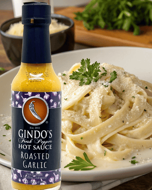 A bottle of Gindo's Fresh Pepper Hot Sauce Roasted Garlic stands in front of a plate of fettuccine pasta garnished with parsley and grated cheese, with a blurred kitchen background.