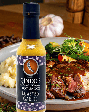 A bottle of Gindo's Fresh Pepper Hot Sauce Roasted Garlic stands in front of a plated meal with sliced steak, mashed potatoes, broccoli, and greens on a wooden table.