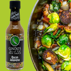 Bottle of Gindo's Spice of Life Hot Sauce Bacon Jalapeno on a green background with a dish of brussels sprouts and bacon.