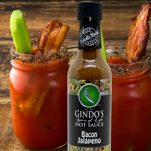Gindo's Bacon Jalapeno hot sauce bottle with Bloody Mary glasses on a wooden background