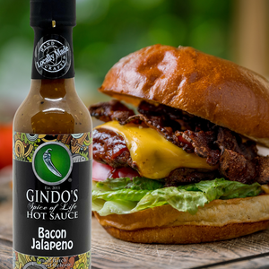 Bottle of Gindo's Bacon Jalapeno hot sauce next to a burger on a wooden surface.