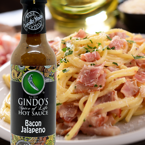 Bottle of Gindo's Spice of Life Hot Sauce Bacon Jalapeno next to a plate of spaghetti pasta carbonara