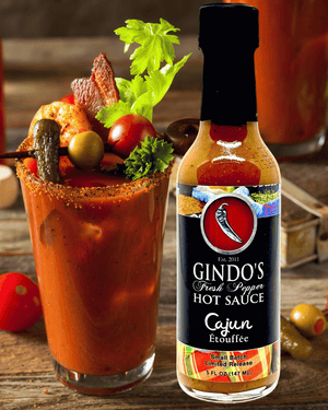 A bottle of Gindo's Fresh Pepper Hot Sauce Cajun Etoufflee stands next to a prepared Bloody Mary cocktail garnished with celery, olives, and bacon on a rustic wooden table.