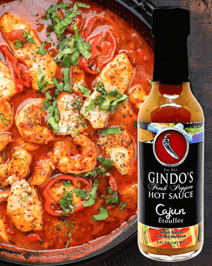 A bottle of Gindo's Cajun Etouffee hot sauce is shown next to a close-up of a shrimp and fish dish cooked in a rich red-orange sauce with parsley garnish, on a dark rustic background.