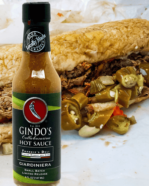 A bottle of Gindo's Collection Collaboration Hot Sauce in the Giardiniera flavor stands in front of an Italian beef sandwich on a white paper liner. The hot sauce bottle has a black cap and a green label with a red logo, while the sandwich behind it contains pulled meat and sliced green jalapeños.