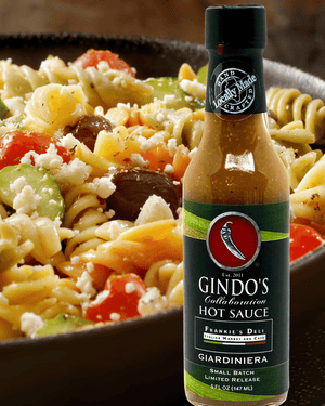 A bottle of Gindo's Giardiniera Hot Sauce placed in front of a close-up photo of a pasta salad with tomatoes, cucumbers, and feta cheese.