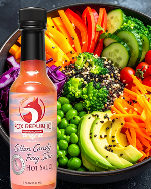 A bottle of Fox Republic Cotton Candy Foxy Sour Hot Sauce is shown in front of a colorful salad bowl filled with sliced avocado, cucumber, broccoli, carrots, peas, and tomatoes on a dark textured background.