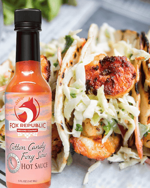 A bottle of Fox Republic Brewing Company Cotton Candy Foxy Saw Hot Sauce placed in front of grilled shrimp tacos filled with shredded coleslaw on a light surface.