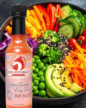 A bottle of Fox Republic Cotton Candy Foxy Sour Hot Sauce stands in front of a large bowl of colorful salad with broccoli, avocado, carrots, peas, cucumber, and tomatoes.